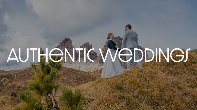 Auf dem Bild steht ein Paar im Allgäu vor den Bergen. Die Braut trägt ein Hochzeitskleid und der Mann seinen Hochzeitsanzug. Davor Beide schauen sich innig an. In weißer Schrift steht Authentic Wedding geschrieben, welches auf die Hochzeitsfotografie von Jan Demann verweist.